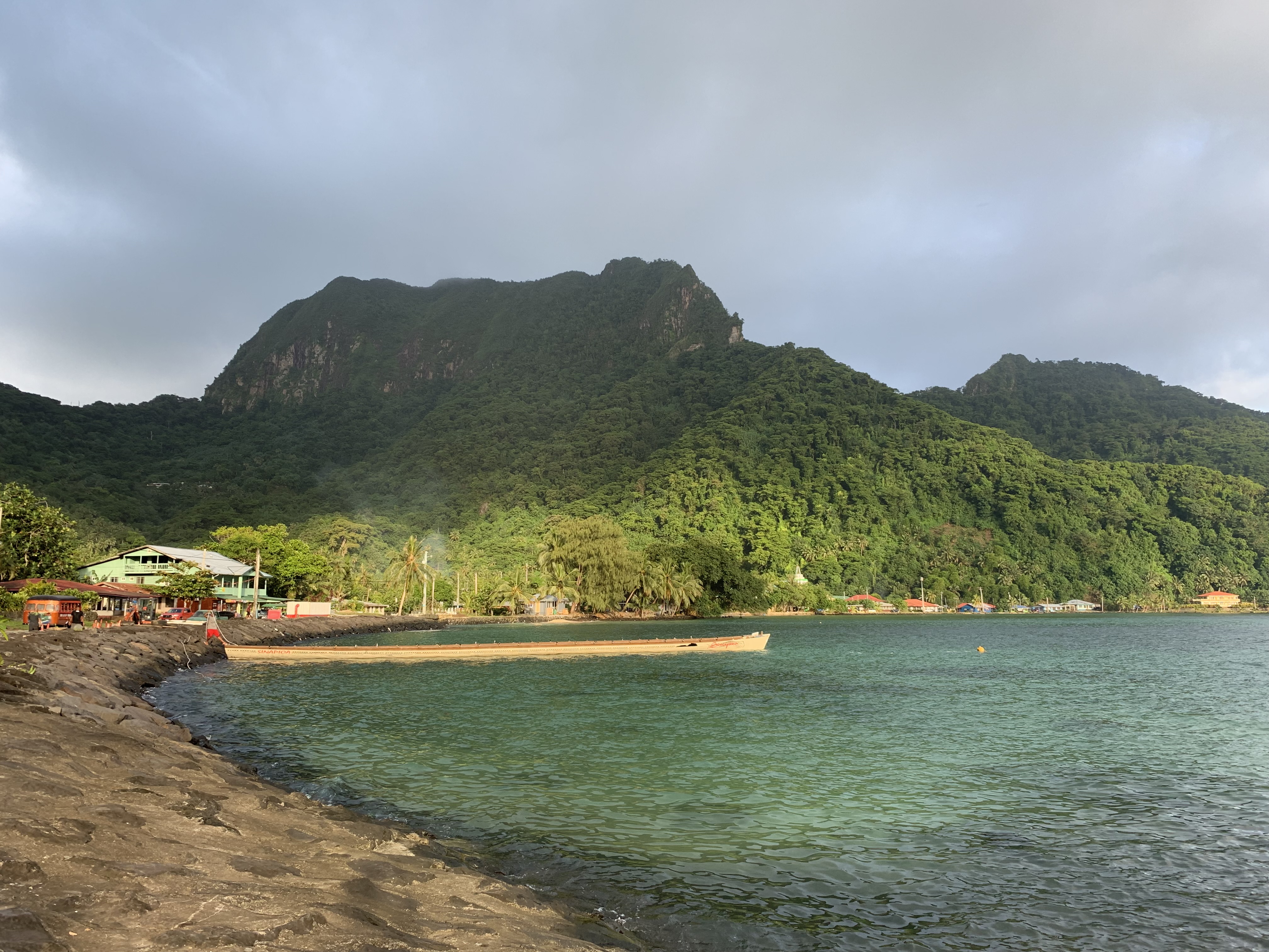 A rocky coast with a large canoe in the water. Tree covered mountains in the distance.