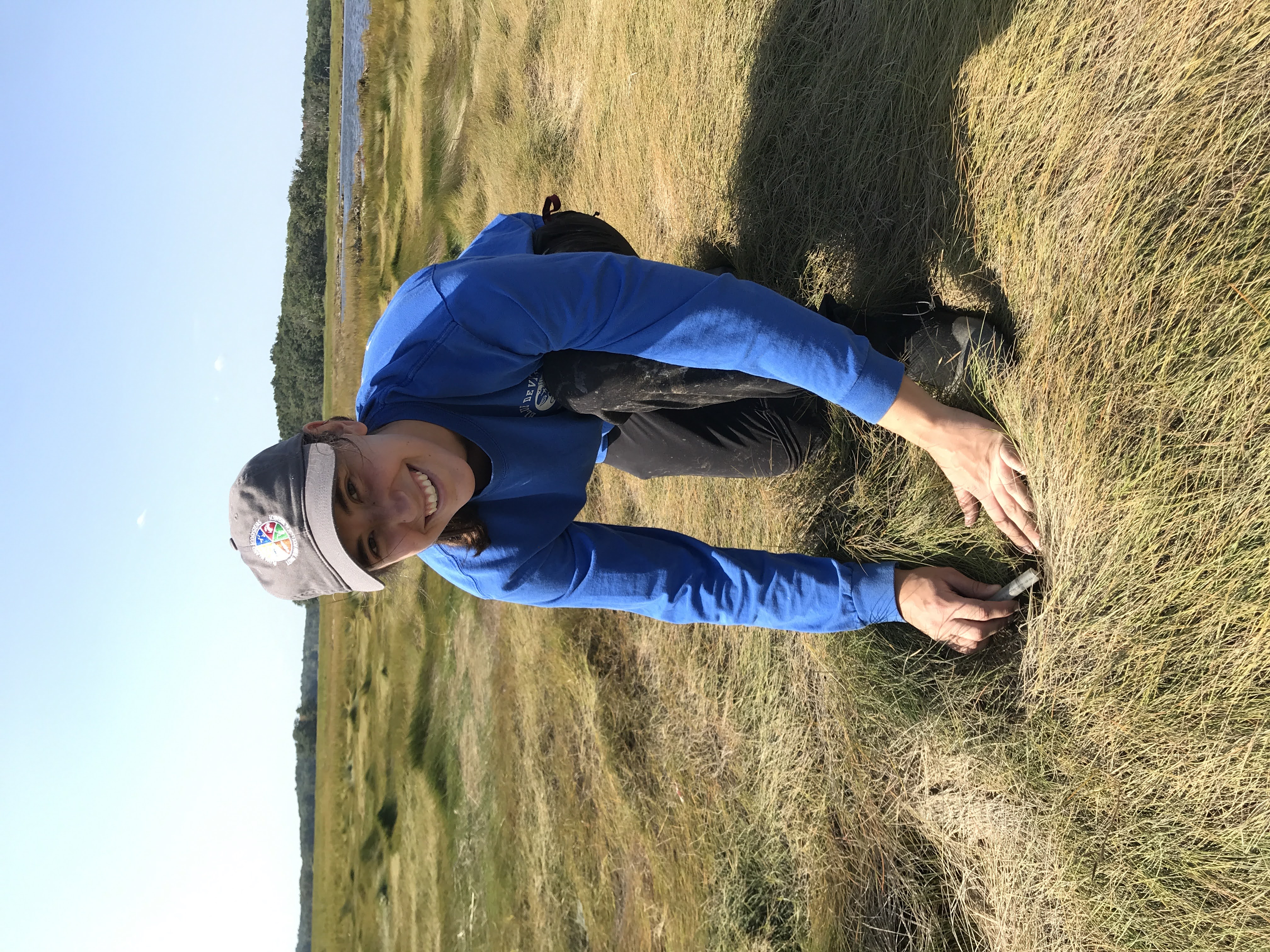 Fellow kneeling and smiling in marsh grass with water and shrubs in the background.