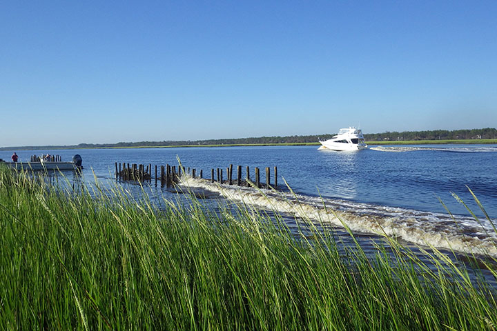 Waves from a traveling yacht roll toward a grassy shoreline protected by a wooden structure.